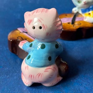 May include: A pink and blue ceramic cat figurine playing a violin. The cat is wearing a blue shirt with white polka dots. The violin is brown with a pink and white string.