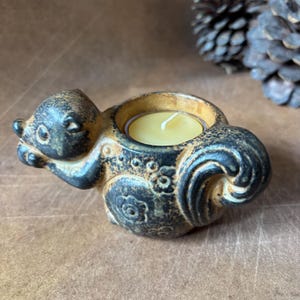 May include: A ceramic squirrel-shaped candle holder in shades of brown and black. The squirrel holds a circular space for a tea light candle. The candle holder has a textured surface and decorative floral patterns. The candle is yellow.