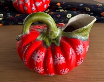 May include: Two ceramic pitchers shaped like red pumpkins with green stems and handles. The pitchers feature a white floral pattern across the ribbed surface and a cream-colored interior. The design mimics the appearance of a gourd with a spout.