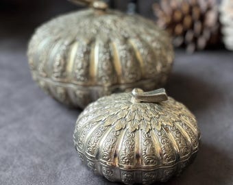May include: Two silver-colored, ornate lidded containers. The containers have a pumpkin-like shape with detailed floral and leaf patterns. The lids have small, rectangular handles. The containers are on a gray surface.