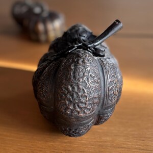 May include: A dark, intricately carved decorative box with a stem-like handle. The box has a pumpkin-like shape and is adorned with detailed floral patterns. The box is made of a dark, possibly metallic material, and sits on a wooden surface.