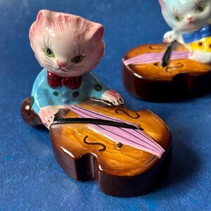 May include: Two ceramic figurines of cats playing the cello. The cat on the left is pink with blue and white polka dot clothing. The cat on the right is white with blue and yellow clothing. Both cats are holding a cello with a brown body and a pink string.