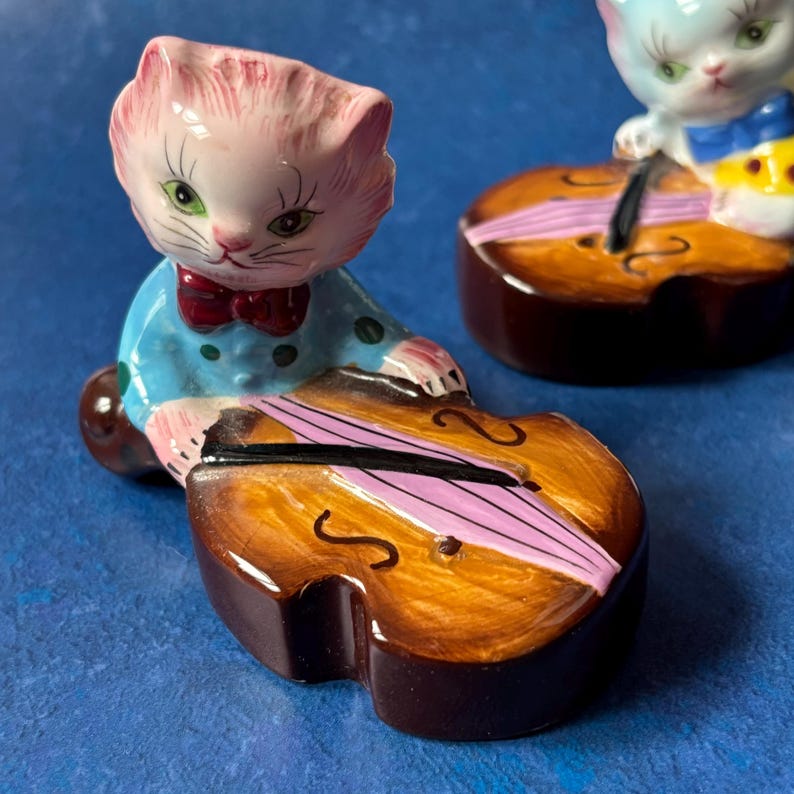 May include: Two ceramic figurines of cats playing the cello. The cat on the left is pink with blue and white polka dot clothing. The cat on the right is white with blue and yellow clothing. Both cats are holding a cello with a brown body and a pink string.