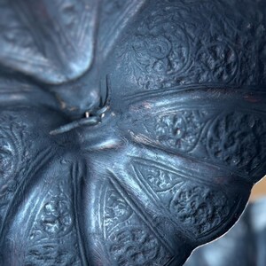 May include: Close-up of a dark, possibly antique, metal object with an intricate floral pattern. The object has a textured surface and appears to be a decorative piece. The color is a deep, muted blue-gray with hints of brown.