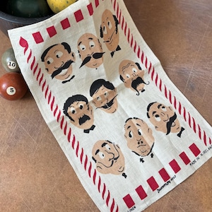 May include: A cream linen tea towel with cartoon faces, each with a different mustache. The towel has a red and white striped border and red square accents. The text "Dunnmore 1960" and "Made in N. Ireland" are printed on the towel.