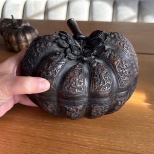 May include: A decorative, dark bronze pumpkin-shaped container with intricate floral carvings. The pumpkin has a stem and decorative accents. Another pumpkin is visible in the background. The container is held by a hand.
