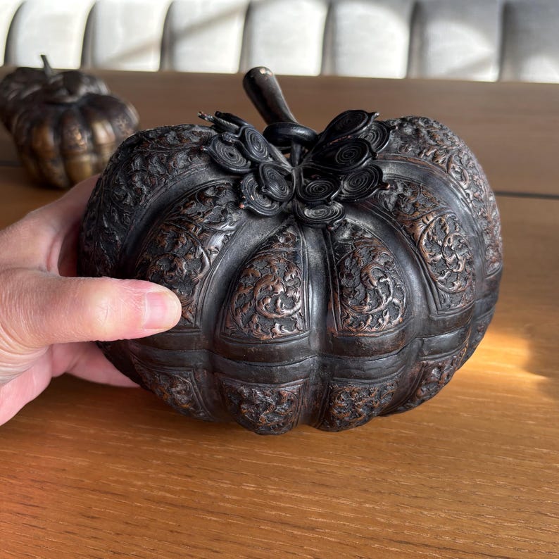 May include: A decorative, dark bronze pumpkin-shaped container with intricate floral carvings. The pumpkin has a stem and decorative accents. Another pumpkin is visible in the background. The container is held by a hand.