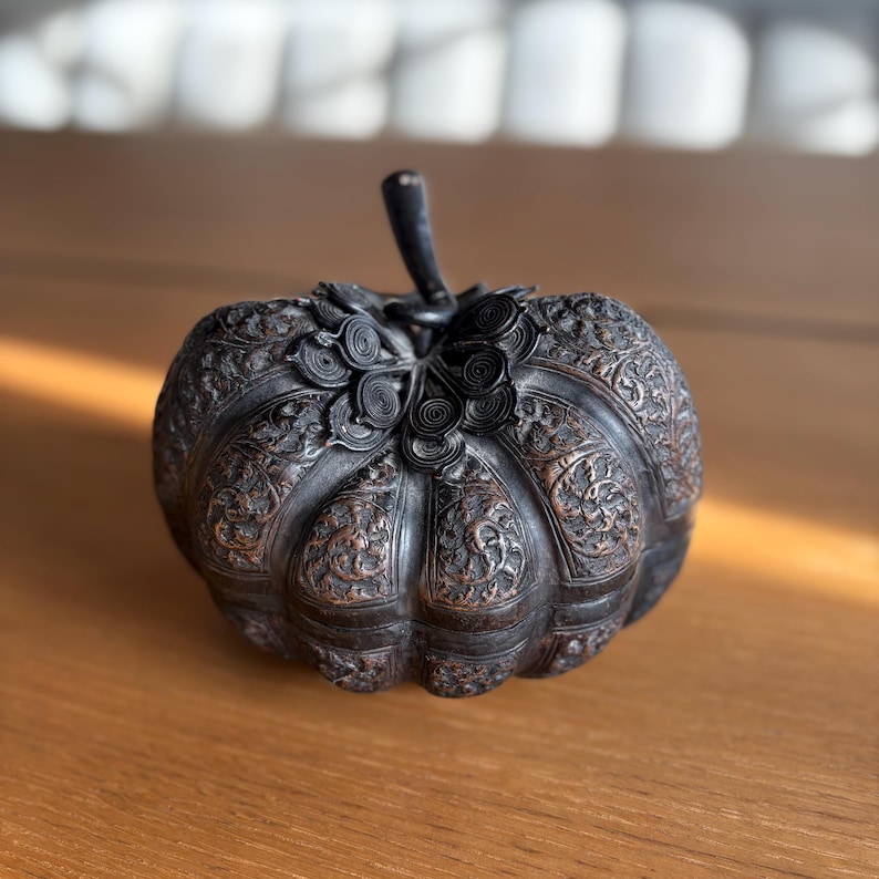 May include: A dark, intricately carved pumpkin-shaped decorative object. The object has a stem and detailed floral patterns. The color is a dark, aged bronze, with a slightly reflective surface. The object is sitting on a wooden surface.
