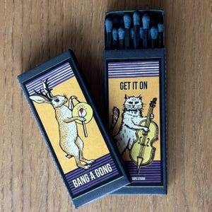 May include: Two matchboxes with illustrations. One features a rabbit playing a gong with the text "Bang a Gong". The other features a cat playing a cello with the text "Get it On".