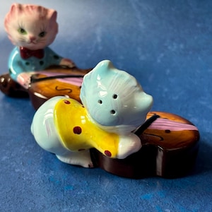 May include: A pair of ceramic cat salt and pepper shakers. The cats are dressed in colorful clothing and are playing a violin. The shakers are in the shape of cats playing a violin.