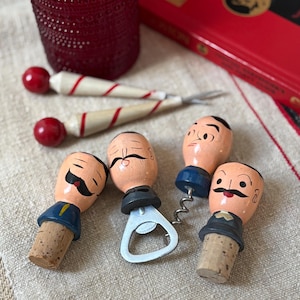 Op de afbeelding: Een verzameling vintage wijnflesstoppers en een flesopener met geschilderde gezichten en snorren. De stoppers hebben kurken bases en zijn beschilderd met cartoonachtige gezichten. Twee rood-wit gestreepte cocktailroerstaafjes zijn ook zichtbaar.