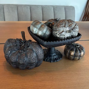 May include: An assortment of decorative metal pumpkins in various sizes and designs. Some are silver-toned, others are bronze, and one is in a black, ornate bowl. The pumpkins have detailed carvings and are arranged on a wooden surface.