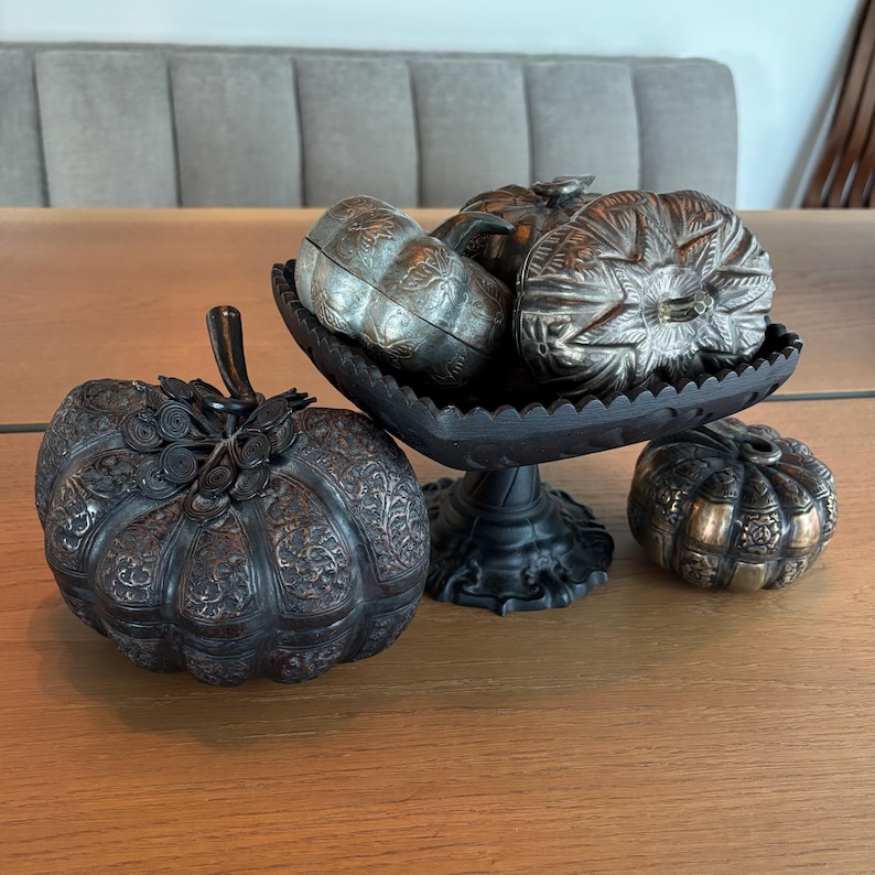 May include: An assortment of decorative metal pumpkins in various sizes and designs. Some are silver-toned, others are bronze, and one is in a black, ornate bowl. The pumpkins have detailed carvings and are arranged on a wooden surface.