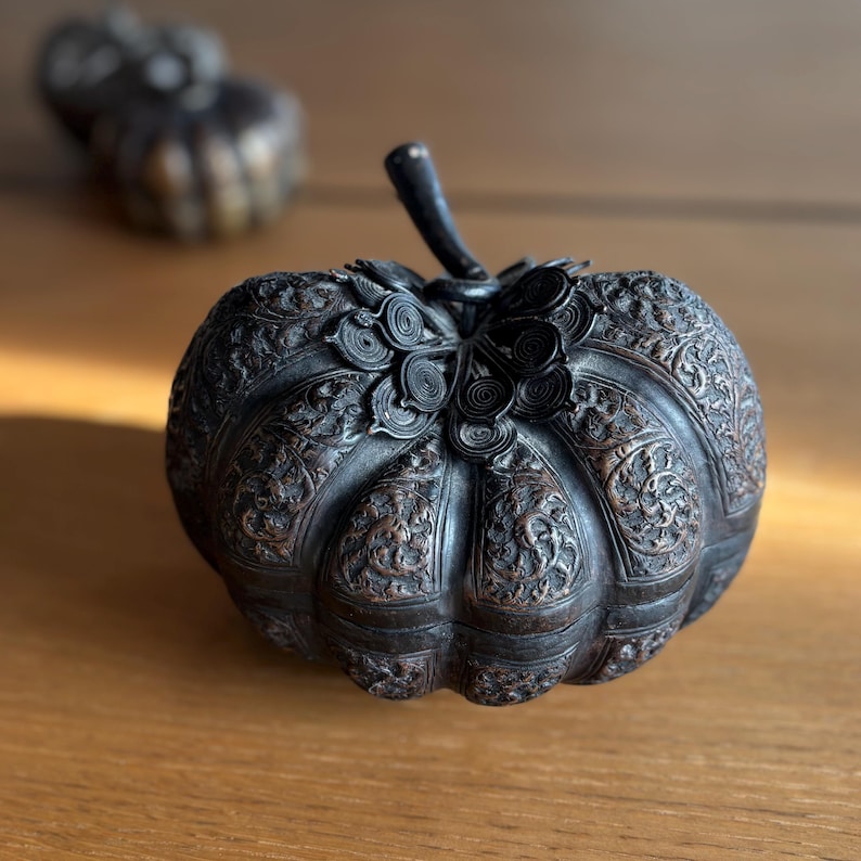 May include: A detailed, dark metal pumpkin sculpture with intricate floral carvings. The pumpkin has a stem and is set on a wooden surface. A second, blurred pumpkin is in the background.