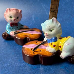 May include: Two ceramic cat figurines playing the violin. The cat on the left is wearing a blue and white outfit with a red bow tie. The cat on the right is wearing a yellow and white outfit with a blue bow tie. Both cats are sitting on top of brown violins.