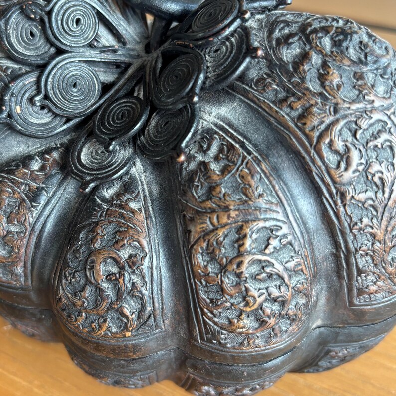 May include: A dark brown, intricately carved decorative box with a pumpkin shape. The box features detailed floral and scrollwork patterns, with a dark, almost black, finish. The lid has a decorative, circular design.