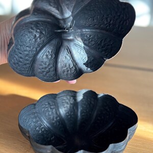 May include: A dark, antique-style metal bowl with a matching lid. The bowl and lid have a scalloped edge and are decorated with intricate floral patterns. The bowl is open, revealing its interior. The lid is slightly raised above the bowl.