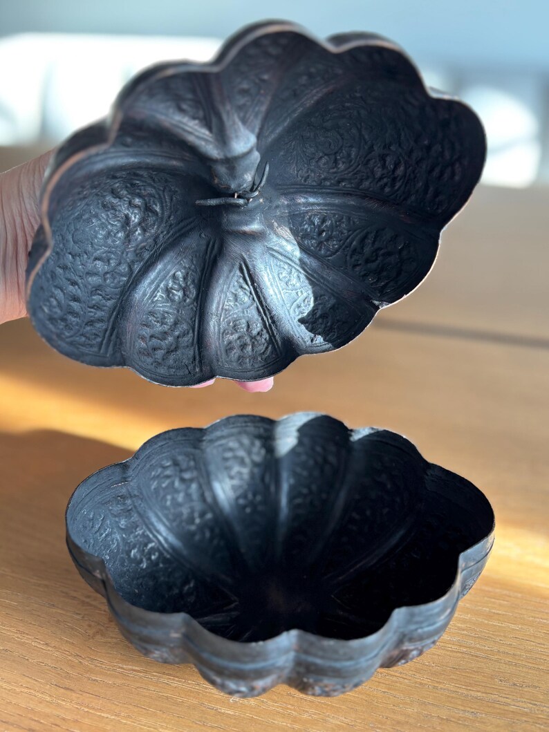 May include: A dark, antique-style metal bowl with a matching lid. The bowl and lid have a scalloped edge and are decorated with intricate floral patterns. The bowl is open, revealing its interior. The lid is slightly raised above the bowl.