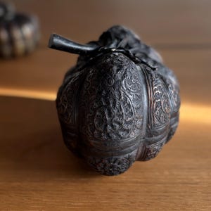May include: A dark, intricately carved metal container, possibly a decorative box or vessel, with a stem-like handle. The surface is covered in detailed floral patterns. The container sits on a wooden surface.