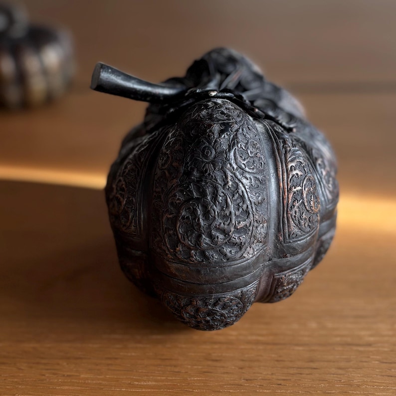 May include: A dark, intricately carved metal container, possibly a decorative box or vessel, with a stem-like handle. The surface is covered in detailed floral patterns. The container sits on a wooden surface.