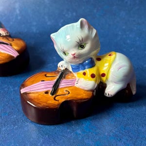 May include: A white ceramic cat figurine playing a cello. The cat is wearing a yellow shirt with red polka dots and a blue bow tie. The cello is brown with pink strings.