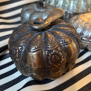 May include: A bronze-colored, pumpkin-shaped container with intricate floral and leaf patterns. The lid features a curved handle. Other similar containers are visible in the background.