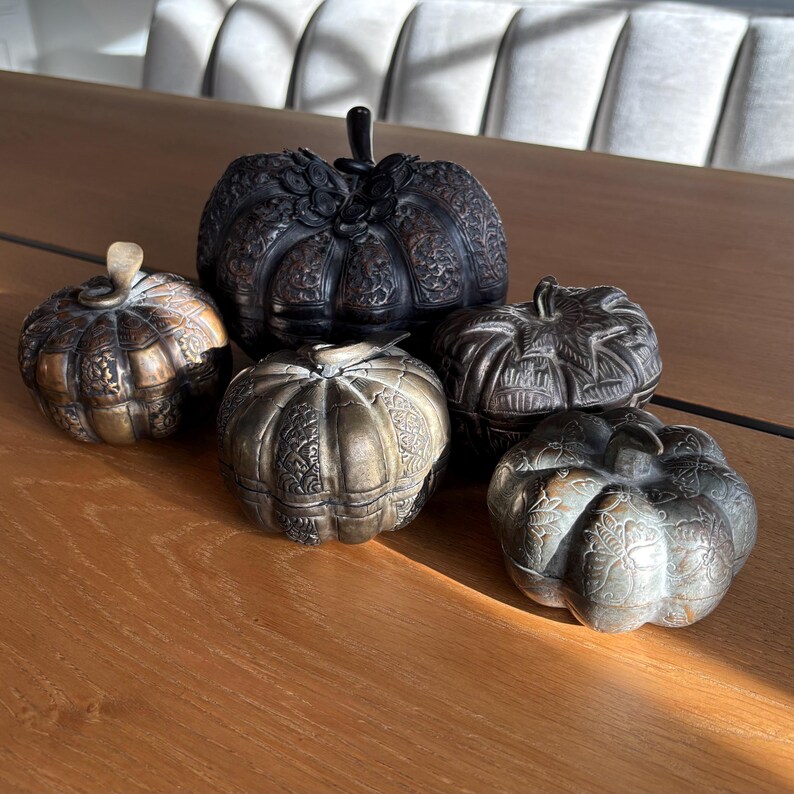 May include: Five decorative pumpkin-shaped containers in varying shades of bronze and black. Each pumpkin has intricate carved details and a stem. The pumpkins are arranged on a wooden surface, suggesting a fall or harvest theme.