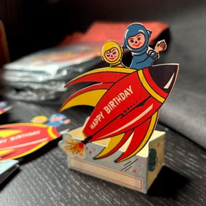 May include: A red and yellow rocket with two astronauts on top. The rocket has the words "Happy Birthday" written on the side. The rocket is a party favor box.