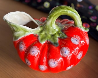 May include: A ceramic creamer shaped like a bright red tomato with a green stem handle and a white interior. The exterior features a repeating pattern of small, light-colored daisy-like flowers with blue centers scattered across the glossy red surface.