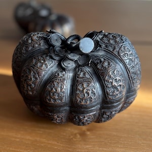 May include: A dark, intricately carved metal container, possibly a box or a decorative piece, shaped like a pumpkin. The surface is covered in detailed floral patterns and scrollwork. The lid features a handle and decorative spirals.