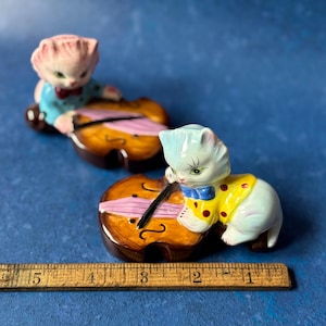 May include: Two ceramic figurines of cats playing the cello. The cat on the left is pink with blue clothing and the cat on the right is blue with a yellow and white polka dot shirt. Both cats are holding a cello with a pink string.