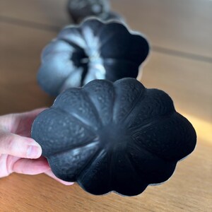 May include: Close-up of three black, decorative, pumpkin-shaped objects. The pumpkins have a textured surface and a scalloped edge. One pumpkin is held in a hand, while the others are in the background. The pumpkins are on a wooden surface.