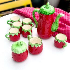 May include: A ceramic strawberry-themed tea set. The set includes a teapot, creamer, sugar bowl, and six teacups. The teapot and cups are red with green accents, while the creamer and sugar bowl are also red with green accents. The set is designed to resemble strawberries.