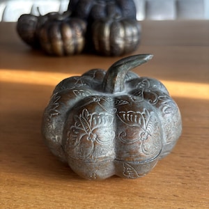 May include: A decorative metal pumpkin with a detailed floral pattern. The pumpkin has a stem and a lid. The color is a mix of bronze and silver. Other pumpkins are visible in the background. The pumpkin is sitting on a wooden surface.