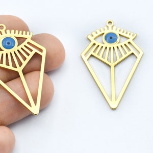 May include: Two gold-colored, geometric pendants in the shape of a diamond, each featuring an eye design with a blue center. The pendants have a small loop at the top for hanging. The eye design is a popular symbol.