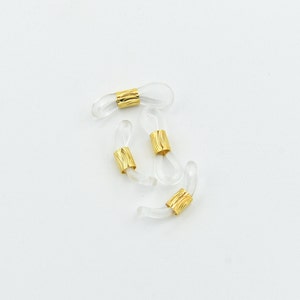 K&ouml;nnte beinhalten: Vier transparente Brillenhalterungen mit goldfarbenen Metallakzenten. Die Halterungen bestehen aus einem transparenten, flexiblen Material und haben eine Schlaufe am Ende, um sie an der Brille zu befestigen. Die goldenen Akzente sind zylindrisch und strukturiert.