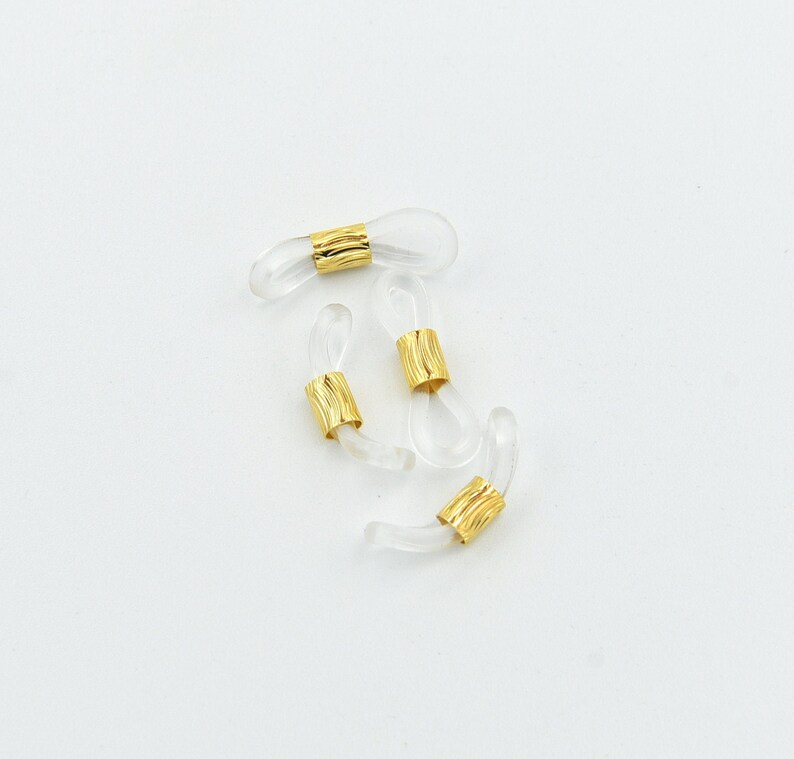 K&ouml;nnte beinhalten: Vier transparente Brillenhalterungen mit goldfarbenen Metallakzenten. Die Halterungen bestehen aus einem transparenten, flexiblen Material und haben eine Schlaufe am Ende, um sie an der Brille zu befestigen. Die goldenen Akzente sind zylindrisch und strukturiert.