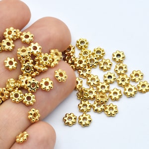 Peut inclure: Un ensemble de petites perles dor&eacute;es en forme de fleur. Chaque perle pr&eacute;sente un trou central et six p&eacute;tales arrondis. Les perles sont dispers&eacute;es sur une surface blanche, sugg&eacute;rant leur utilisation en bijouterie ou en artisanat.