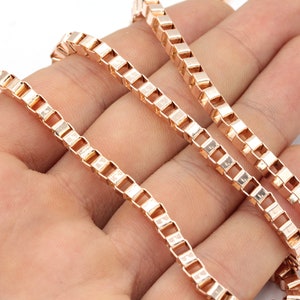 May include: Rose gold-colored box chain necklace. The chain is made of small, square links that create a smooth, continuous surface. The necklace is displayed against a neutral background, highlighting its metallic sheen.