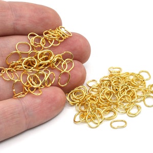 May include: A collection of small, gold-coloured oval-shaped jewellery findings. The findings are scattered on a white surface, with some held in a hand. These findings are likely used for jewellery making and crafting.