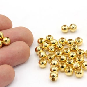 May include: A collection of small, shiny, gold-colored metal beads. The beads are round with a hole through the center, and are scattered on a white surface. Some beads are held in a person's hand.