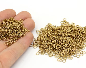 Puede incluir: Una colección de pequeños anillos de salto dorados, dispersos sobre una superficie blanca. Algunos anillos se sostienen en una mano, mientras que otros se apilan en un grupo más grande. Estos anillos metálicos se utilizan para la fabricación de joyas y otras manualidades.