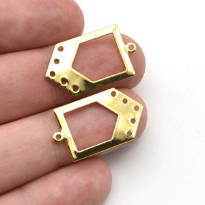 Könnte beinhalten: Zwei goldfarbene Metall-Schmuckkomponenten. Jede Komponente hat eine geometrische Form mit einem zentralen offenen Raum und mehreren kleinen Löchern. Sie haben auch eine kleine Schlaufe zur Befestigung. Die Komponenten befinden sich auf weißem Hintergrund.