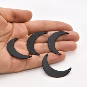 May include: Five black crescent moon charms with small holes at each end. These metal charms are ideal for jewelry making or other craft projects. The charms are held in a hand against a white background.