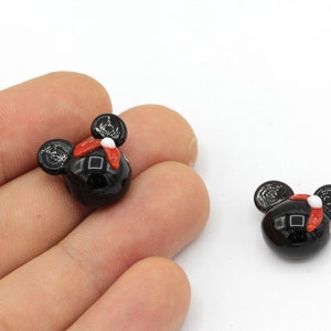 Puede incluir: Dos cuentas negras con forma de Mickey Mouse con detalles rojos y blancos. Las cuentas tienen un acabado brillante sobre un fondo blanco. Las cuentas son peque&ntilde;as y parecen estar hechas de vidrio o un material similar.