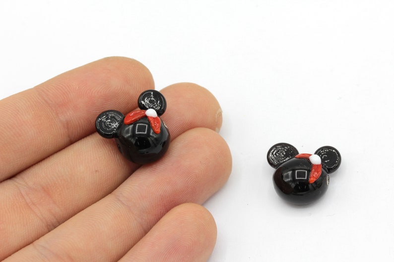 Puede incluir: Dos cuentas negras con forma de Mickey Mouse con detalles rojos y blancos. Las cuentas tienen un acabado brillante sobre un fondo blanco. Las cuentas son peque&ntilde;as y parecen estar hechas de vidrio o un material similar.