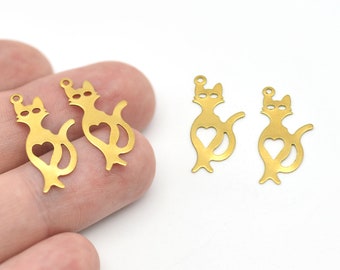 Puede incluir: Encantos dorados en forma de gato con un corazón recortado en el centro. Los amuletos tienen un pequeño lazo en la parte superior para sujetarlos a las joyas. Los amuletos de gato son de metal y se muestran sobre un fondo blanco.