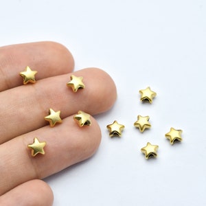 May include: A collection of small, gold-colored star-shaped beads. The beads are scattered on a white surface, with some held by a person's fingers. The stars have a matte finish and a five-point design, perfect for jewelry making or crafts.