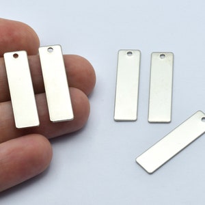 May include: Several rectangular, silver-colored metal tags with a small hole at the top. Two tags are held in a hand, while the others are scattered on a white surface. These tags are suitable for jewelry making or crafting.