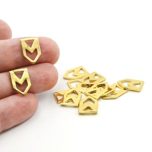 Könnte beinhalten: Goldfarbene Metallanhänger in Schildform mit einem ausgeschnittenen Chevron-Design. Die Anhänger sind klein, mit poliertem Finish, und liegen auf einer weißen Oberfläche verstreut. Einige werden von einer Hand gehalten.